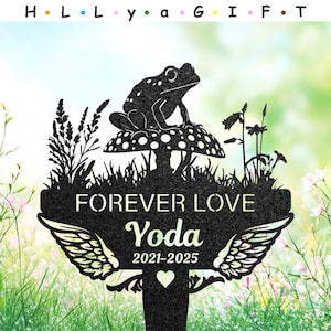 Puede incluir: Una estaca de jardín de metal negro con una rana sobre una seta. La estaca dice "FOREVER LOVE" con el nombre "Yoda" y las fechas "2021-2025". El diseño incluye detalles de alas y un corazón, sobre un fondo de jardín borroso.