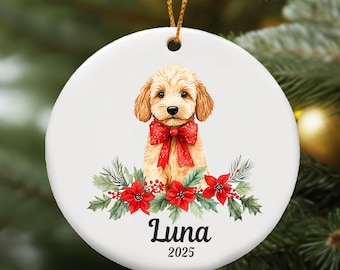 Puede incluir: Adorno navideño redondo de cerámica blanca con un perro golden doodle con un lazo rojo. El adorno está decorado con flores de pascua rojas, follaje verde y el nombre "Luna" con el año "2025" en texto negro.