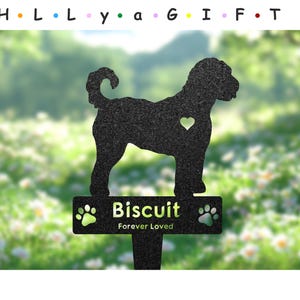 Op de afbeelding: Zwarte metalen silhouet van een hond met een hart op zijn borst en de woorden "Biscuit Forever Loved" op een plaquette eronder. De silhouet staat op een groene achtergrond met witte bloemen.