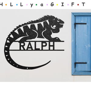 May include: Black metal silhouette of an iguana with a curved tail and the name "RALPH" written on a banner below. The iguana is facing right and has a textured surface.