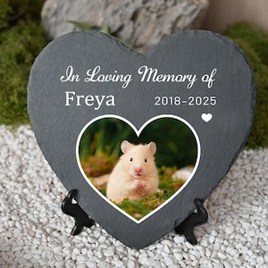 May include: A heart-shaped slate memorial plaque with the text "In Loving Memory of Freya 2018-2025." A photo of a hamster is in the center. The plaque is supported by a black stand and rests on white pebbles.