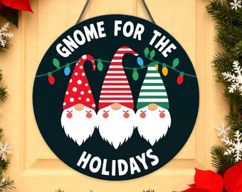 May include: Round black wooden sign with the text "Gnome for the Holidays" and three gnome illustrations. The gnomes wear red, white, and green hats and have white beards. A string of colorful lights is above the gnomes.