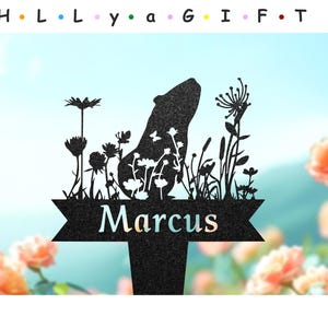 May include: Black metal silhouette of a guinea pig with a name sign that reads "Marcus" and a floral design. The silhouette is set against a blue sky background.