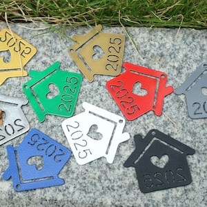 K&ouml;nnte beinhalten: Eine Sammlung von acht hausf&ouml;rmigen Ornamenten in verschiedenen Farben, darunter Gold, Gr&uuml;n, Rot, Wei&szlig;, Grau und Blau. Jedes Ornament zeigt ein Herz und das Jahr 2025.