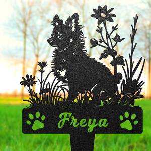 K&ouml;nnte beinhalten: Schwarze Metallsilhouette eines Hundes mit Blumen und dem Namen "Freya" auf einer Plakette. Die Silhouette ist vor einem verschwommenen Hintergrund aus gr&uuml;nem Gras und B&auml;umen platziert.