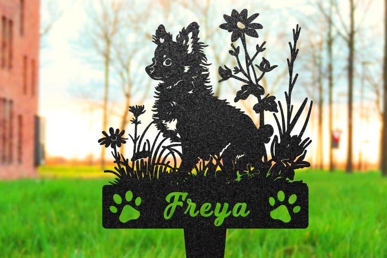K&ouml;nnte beinhalten: Schwarze Metallsilhouette eines Hundes mit Blumen und dem Namen "Freya" auf einer Plakette. Die Silhouette ist vor einem verschwommenen Hintergrund aus gr&uuml;nem Gras und B&auml;umen platziert.