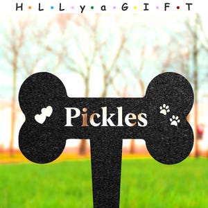 May include: Black bone-shaped garden stake with the name "Pickles" in white and copper lettering. Features two white hearts on the left and two paw prints on the right. The text "H L L y a G I F T" is above the stake.