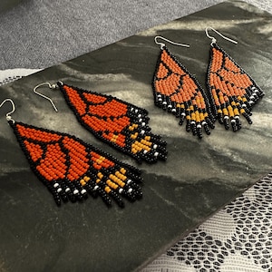 Può includere: Un paio di orecchini pendenti con un design a farfalla monarca. Gli orecchini sono realizzati con perline nere, arancioni e gialle.
