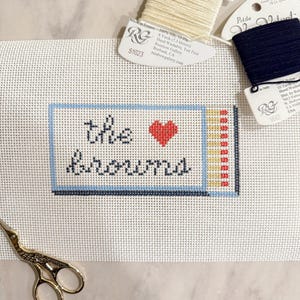 May include: A white canvas with a blue and yellow border, a red heart, and the text "the rumors" in black. The canvas is for needlepoint and cross stitch.