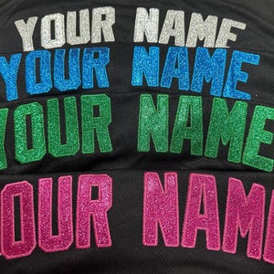K&ouml;nnte beinhalten: Eine schwarze Stoffoberfl&auml;che mit vier Zeilen des Textes YOUR NAME in glitzernden Blockbuchstaben. Die oberste Reihe ist silber, die zweite blau, die dritte gr&uuml;n und die untere Reihe pink. Jeder Buchstabe weist eine texturierte Oberfl&auml;che auf.