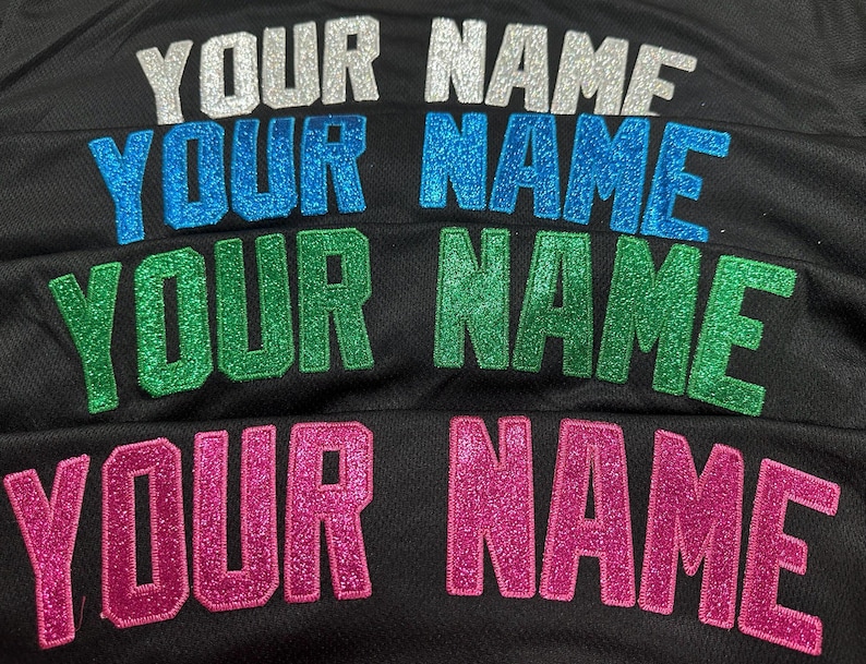 K&ouml;nnte beinhalten: Eine schwarze Stoffoberfl&auml;che mit vier Zeilen des Textes YOUR NAME in glitzernden Blockbuchstaben. Die oberste Reihe ist silber, die zweite blau, die dritte gr&uuml;n und die untere Reihe pink. Jeder Buchstabe weist eine texturierte Oberfl&auml;che auf.