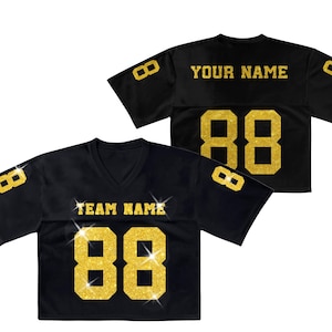 K&ouml;nnte beinhalten: Ein schwarzes kurz&auml;rmeliges Trikot mit goldfarbenen Glitzerbuchstaben und -zahlen. Auf der Vorderseite steht TEAM NAME &uuml;ber der Nummer 88, auf der R&uuml;ckseite YOUR NAME &uuml;ber der Nummer 88. Weitere 8er befinden sich auf den &Auml;rmeln.
