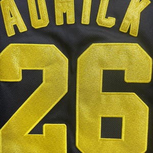 May include: Close-up of a navy blue jersey with gold embroidered lettering and numbers. The name "AUMICK" is arched across the top, with the number "26" below. The jersey has a mesh texture.