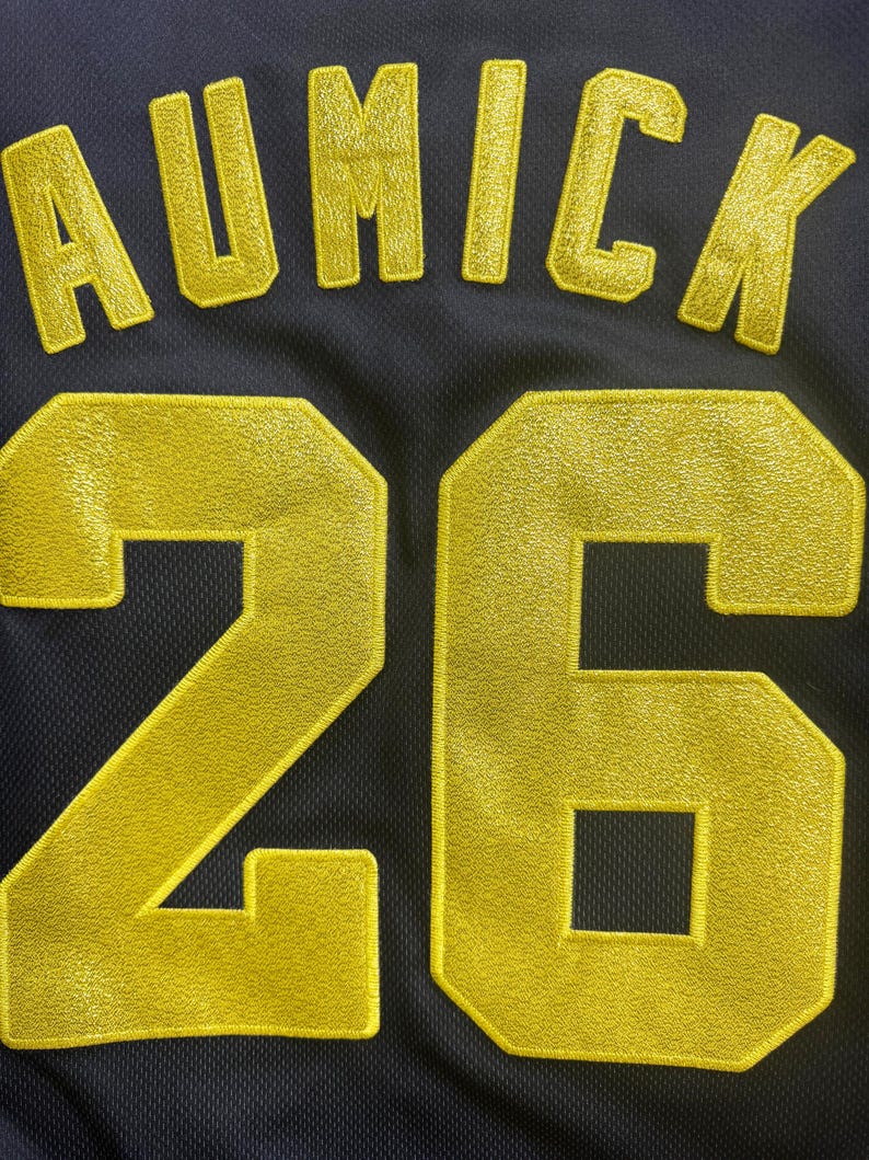 May include: Close-up of a navy blue jersey with gold embroidered lettering and numbers. The name "AUMICK" is arched across the top, with the number "26" below. The jersey has a mesh texture.