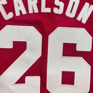 K&ouml;nnte beinhalten: Ein rotes Sporttrikot mit dem Namen CARLSON in wei&szlig;en Blockbuchstaben, die oben bogenf&ouml;rmig angeordnet sind. Unter dem Namen ist die Nummer 26 in gro&szlig;en, wei&szlig;en, aufgen&auml;hten Blockziffern mit einer leicht schimmernden Textur zu sehen.