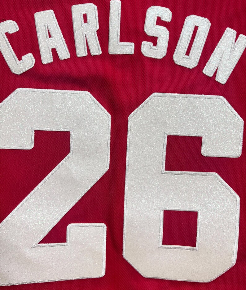 K&ouml;nnte beinhalten: Ein rotes Sporttrikot mit dem Namen CARLSON in wei&szlig;en Blockbuchstaben, die oben bogenf&ouml;rmig angeordnet sind. Unter dem Namen ist die Nummer 26 in gro&szlig;en, wei&szlig;en, aufgen&auml;hten Blockziffern mit einer leicht schimmernden Textur zu sehen.