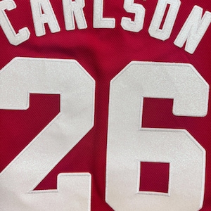 May include: Close-up of a red jersey with the name "CARLSON" in white letters above the number "26". The numbers and letters are outlined in white and stitched onto the jersey.