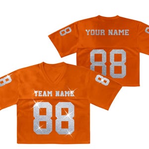 K&ouml;nnte beinhalten: Zwei orangefarbene V-Ausschnitt-Trikots mit kurzen &Auml;rmeln. Die Vorderseite zeigt den Text TEAM NAME und die Nummer 88 in silbernem Glitzer. Die R&uuml;ckseite zeigt YOUR NAME und die Nummer 88. Beide &Auml;rmel haben die Nummer 8 in silbernem Glitzer.