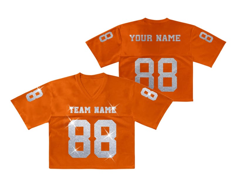 K&ouml;nnte beinhalten: Zwei orangefarbene V-Ausschnitt-Trikots mit kurzen &Auml;rmeln. Die Vorderseite zeigt den Text TEAM NAME und die Nummer 88 in silbernem Glitzer. Die R&uuml;ckseite zeigt YOUR NAME und die Nummer 88. Beide &Auml;rmel haben die Nummer 8 in silbernem Glitzer.