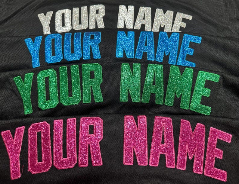 May include: Close-up of a black mesh fabric with the words "YOUR NAME" embroidered in silver, blue, green, and pink glitter. The text is in a bold, blocky font. The image showcases personalized apparel.