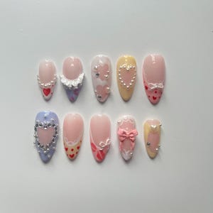 May include: Ten different false nail designs with various embellishments, including hearts, bows, and pearls. The nails are all almond shaped and have a light pink base.