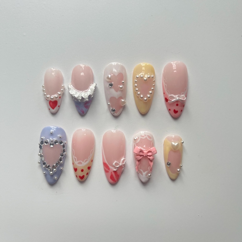 May include: Ten different false nail designs with various embellishments, including hearts, bows, and pearls. The nails are all almond shaped and have a light pink base.