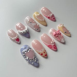 May include: Set of ten false nails with different designs. Each nail is decorated with a heart, pearls, a bow, or lace. The nails are in various colors, including pink, yellow, blue, and white.