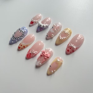 May include: Set of ten pink, yellow, and blue false nails with various designs, including hearts, bows, and lace.