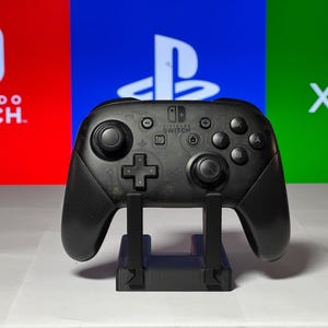 May include: A black Nintendo Switch Pro Controller on a stand in front of a red, blue, and green background with the logos for Nintendo Switch, Playstation, and Xbox.