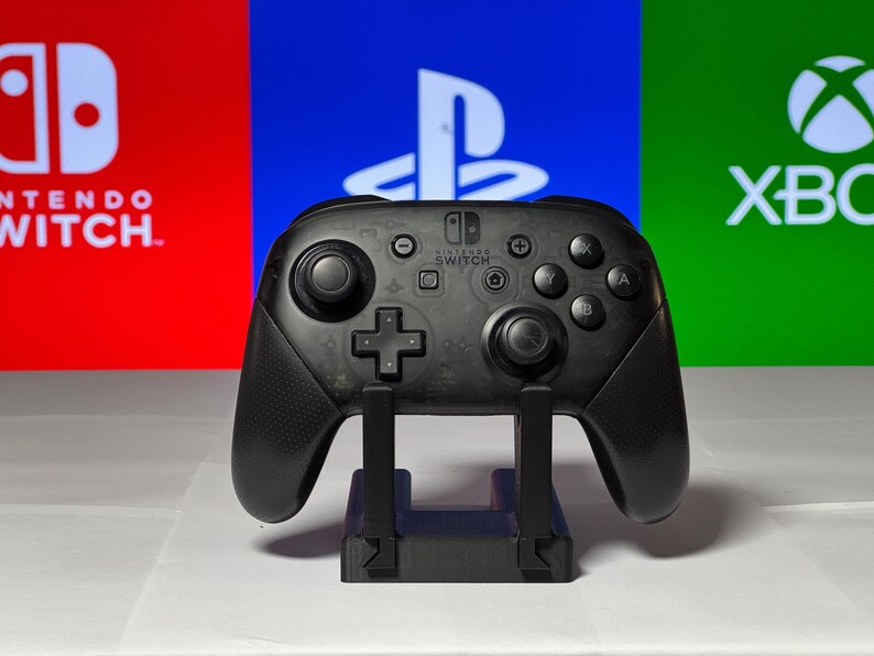 May include: A black Nintendo Switch Pro Controller on a stand in front of a red, blue, and green background with the logos for Nintendo Switch, Playstation, and Xbox.