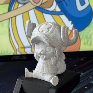 以下が含まれることがあります： 大きな帽子と角を持つ漫画のキャラクターの小さな灰色のフィギュア。黒い台座に座っています。フィギュアはテクスチャ加工された表面で、背景はぼやけています。