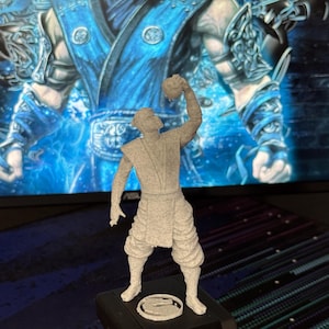 Może przedstawiać: Szara figurka 3D postaci z Mortal Kombat. Figurka stoi na czarnej podstawie z okrągłym emblematem. Tło przedstawia niebiesko-biały obraz.