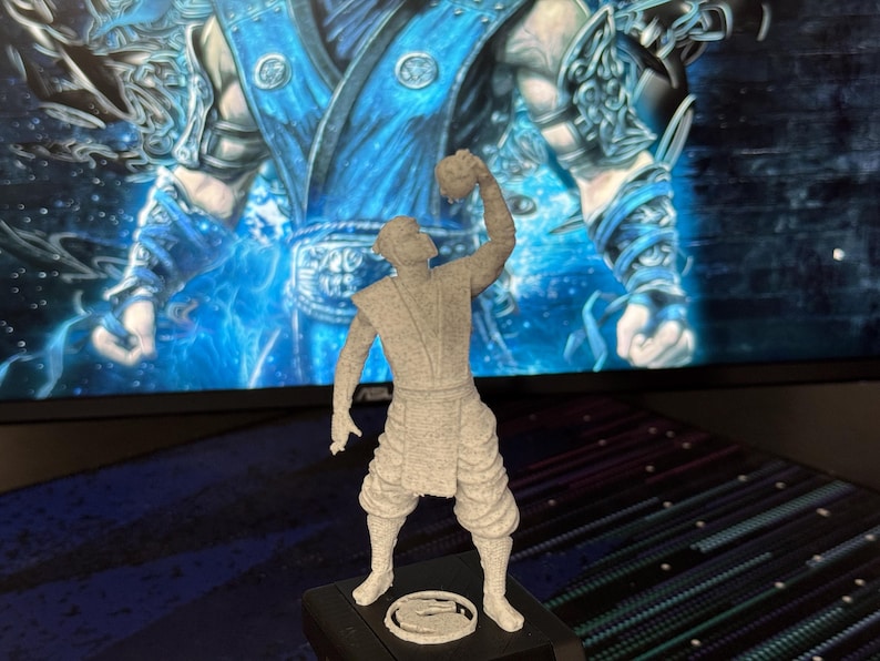 Może przedstawiać: Szara figurka 3D postaci z Mortal Kombat. Figurka stoi na czarnej podstawie z okrągłym emblematem. Tło przedstawia niebiesko-biały obraz.