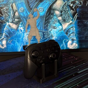 Może przedstawiać: Czarny kontroler Nintendo Switch Pro w czarnym stojaku, z małą szarą figurką postaci z gry wideo na g&oacute;rze. Tło to monitor komputera wyświetlający niebiesko-białą grafikę.