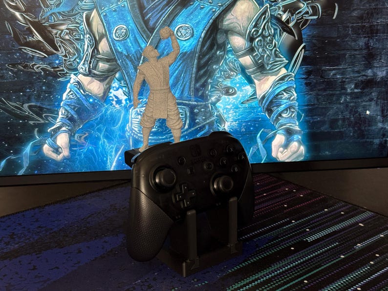 Może przedstawiać: Czarny kontroler Nintendo Switch Pro w czarnym stojaku, z małą szarą figurką postaci z gry wideo na g&oacute;rze. Tło to monitor komputera wyświetlający niebiesko-białą grafikę.