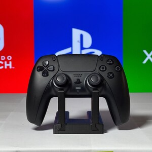 May include: A black PlayStation 5 DualSense controller is displayed on a stand against a backdrop of red, blue, and green screens with the logos for Nintendo Switch, PlayStation, and Xbox.
