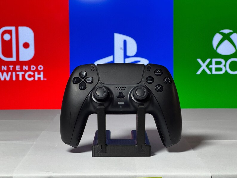 May include: A black PlayStation 5 DualSense controller is displayed on a stand against a backdrop of red, blue, and green screens with the logos for Nintendo Switch, PlayStation, and Xbox.
