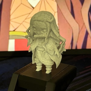 Puede incluir: Una pequeña figura de color claro de un personaje con pelo largo y un atuendo distintivo. La figura está sobre una superficie negra, con un fondo colorido visible detrás. La figura tiene una textura detallada.