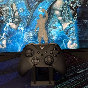 Może przedstawiać: Szara figurka z nadrukiem 3D postaci z gry wideo stoi na czarnym kontrolerze Xbox. Kontroler spoczywa na czarnym stojaku. Tło to monitor komputera wyświetlający niebiesko-białą grafikę.