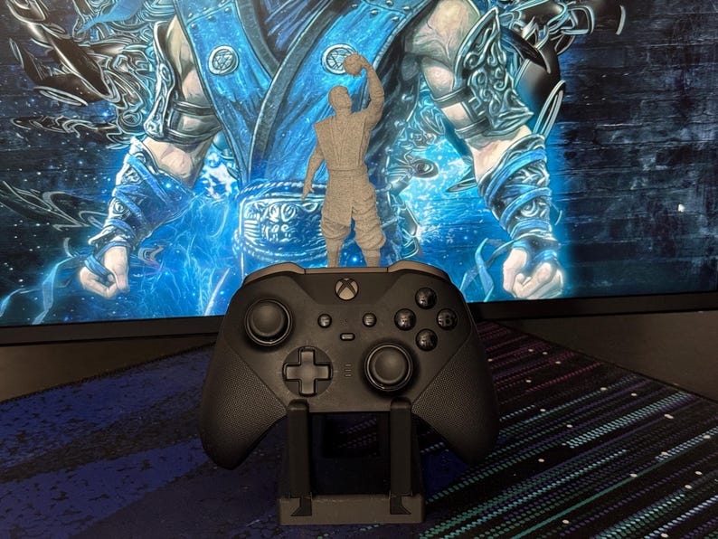 Może przedstawiać: Szara figurka z nadrukiem 3D postaci z gry wideo stoi na czarnym kontrolerze Xbox. Kontroler spoczywa na czarnym stojaku. Tło to monitor komputera wyświetlający niebiesko-białą grafikę.