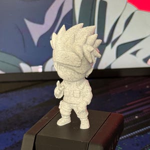May include: A small, light gray figurine of a character with spiky hair and a blindfold. The figure is in a standing pose, with one hand raised in a peace sign. The figurine is on a black surface, with a blurred background.
