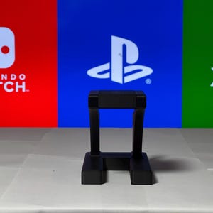 May include: A black plastic stand for a video game console. The stand is in front of a background with the logos for Nintendo Switch, PlayStation, and Xbox.