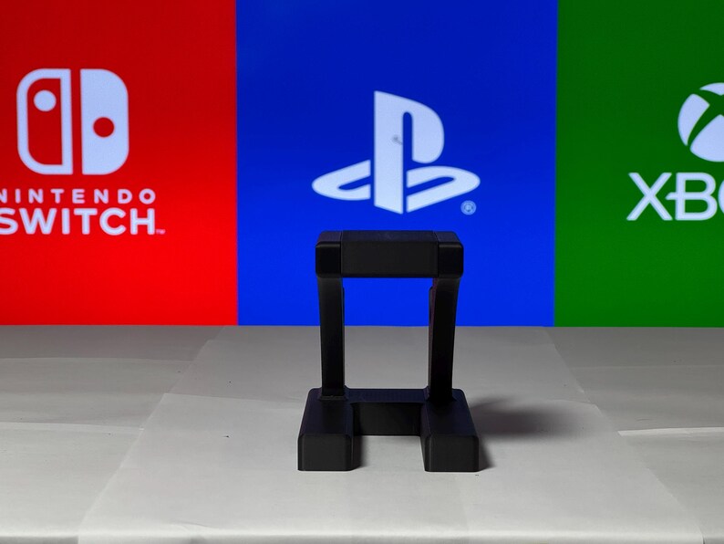 May include: A black plastic stand for a video game console. The stand is in front of a background with the logos for Nintendo Switch, PlayStation, and Xbox.