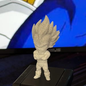 Puede incluir: Una pequeña figura de color claro de un personaje con pelo puntiagudo y los brazos cruzados. La figura está sobre una base negra, con un fondo borroso con colores azules y blancos. La figura parece ser un objeto de colección.