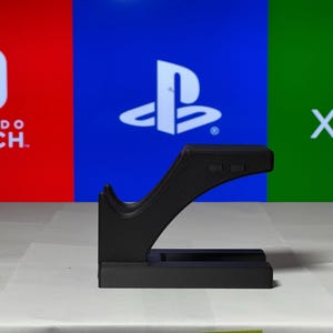 May include: Black plastic stand for a video game controller. The stand has a curved design and a flat base. The stand is in front of a background with the logos for Nintendo Switch, Playstation, and Xbox.