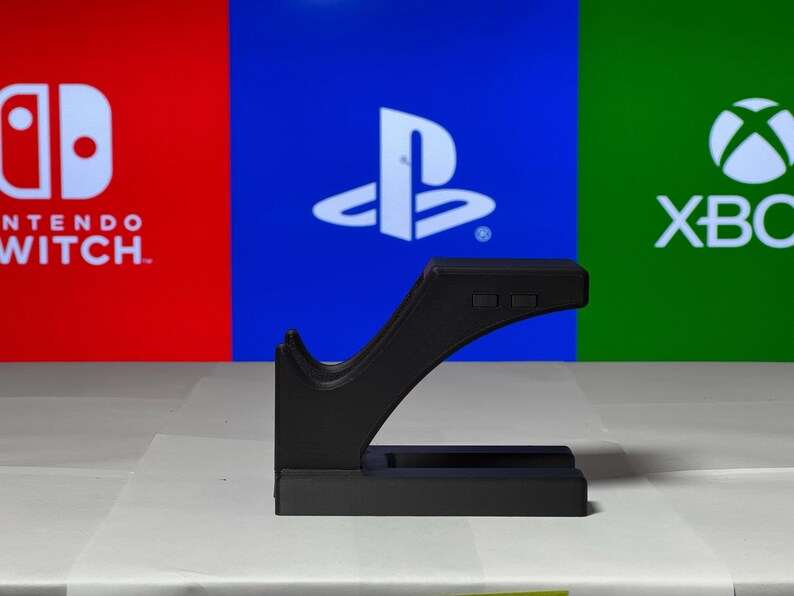 May include: Black plastic stand for a video game controller. The stand has a curved design and a flat base. The stand is in front of a background with the logos for Nintendo Switch, Playstation, and Xbox.