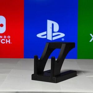 May include: A black plastic stand for holding a video game controller. The stand is in front of a background with the logos for Nintendo Switch, PlayStation, and Xbox.
