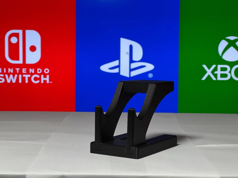 May include: A black plastic stand for holding a video game controller. The stand is in front of a background with the logos for Nintendo Switch, PlayStation, and Xbox.
