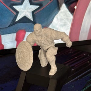 Könnte beinhalten: Eine 3D-gedruckte Captain America-Figur in kniender Pose, die ihren Schild hält. Die Figur ist hellgrau gesprenkelt und auf einem schwarzen Sockel montiert. Der Hintergrund zeigt ein Captain America-Bild.