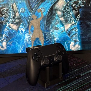 Może przedstawiać: Czarny kontroler PlayStation 5 spoczywający na czarnym stojaku, z małą figurką postaci z gry wideo na g&oacute;rze. Tło przedstawia niebiesko-biały obraz sztuki cyfrowej.